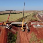 Vista aérea de uma ponte em construção, com guindastes e máquinas no canteiro de obras.